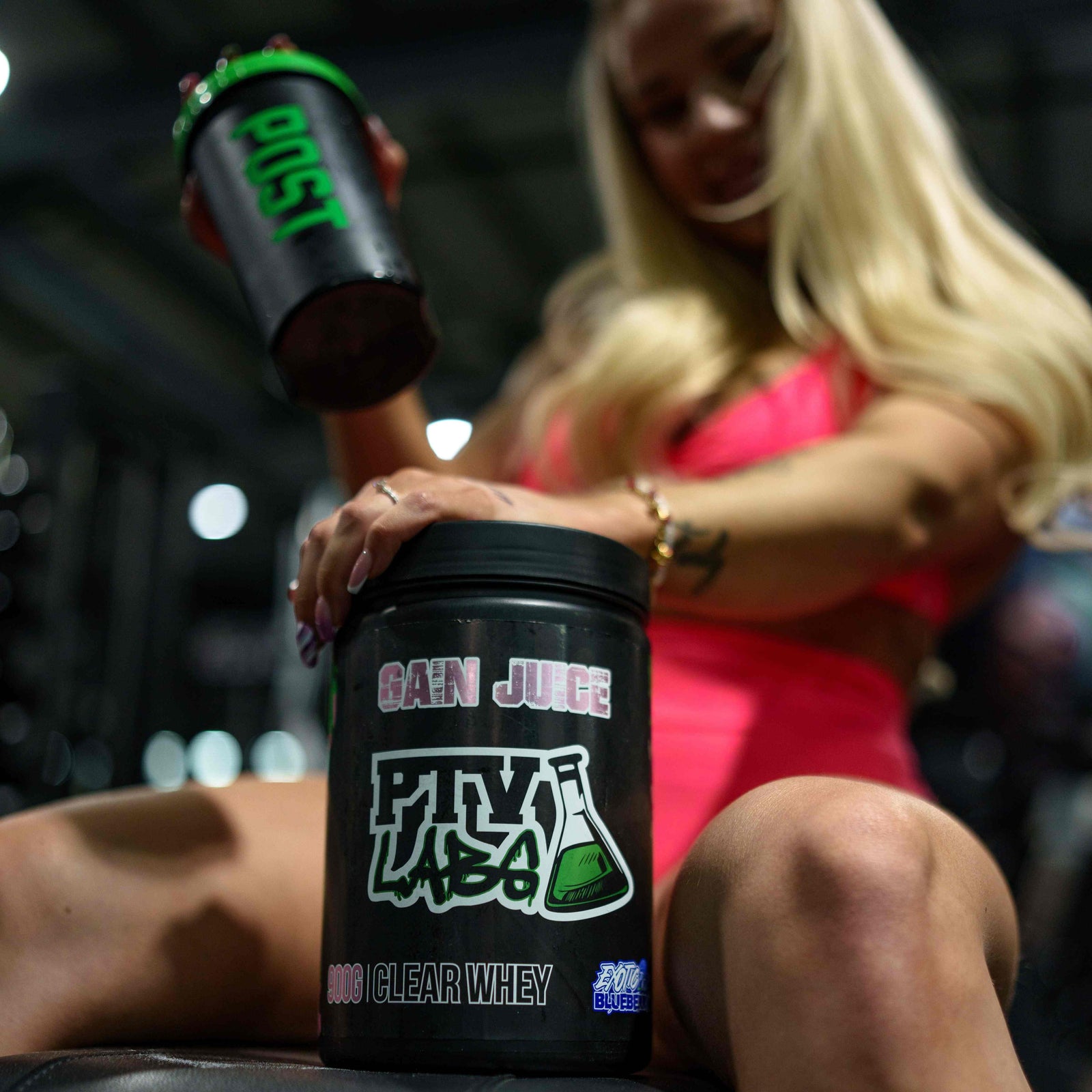 Nainen kuntosalilla pitää kädessään PTVLABS Gain Juice -lisäravinnejuomaa ja esittelee PTVLABS Clear Whey -proteiinituotetta.