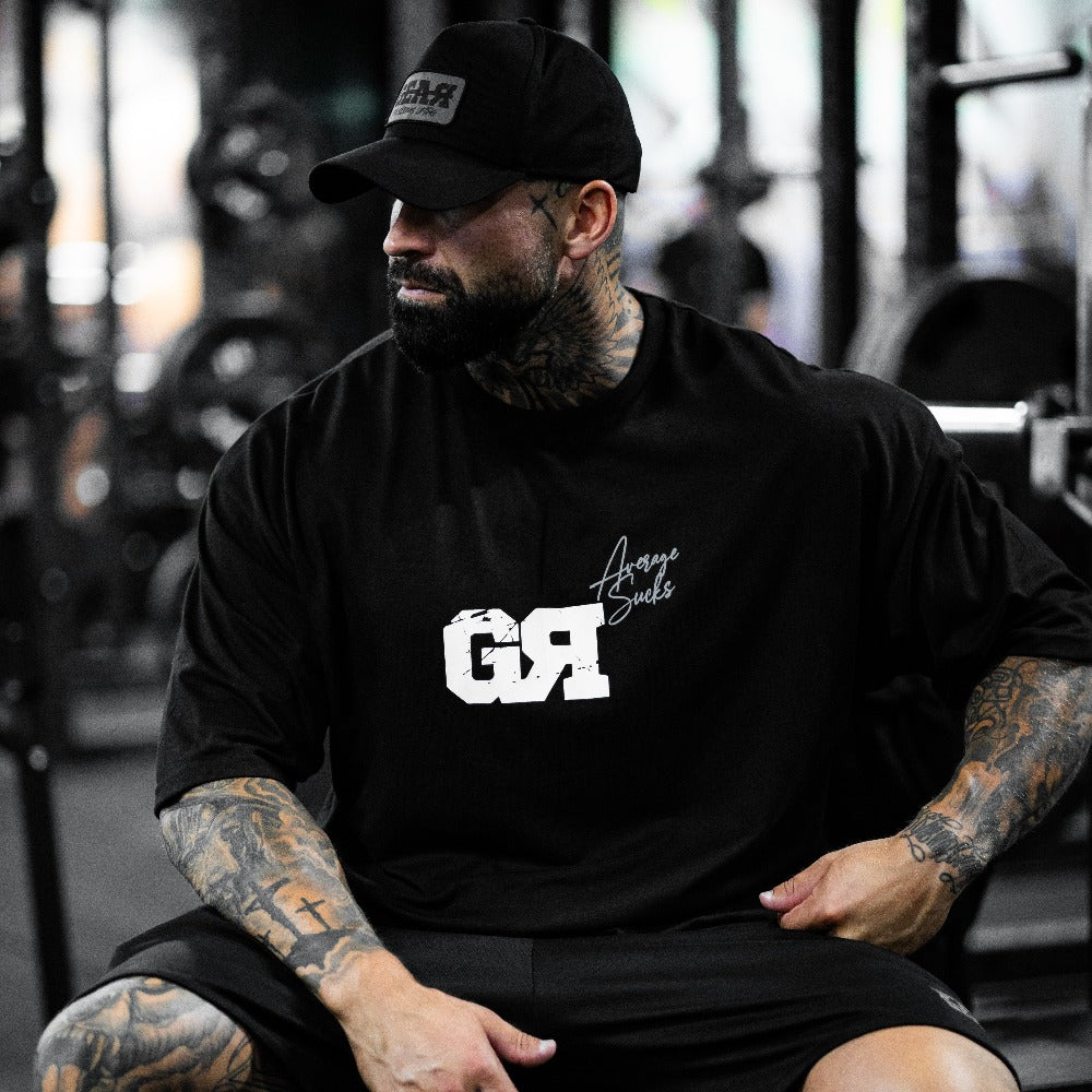 Oversized musta treenipaita salille – GR-logo ja 'Average Sucks' -teksti, miesten urheilupaita rennolla istuvuudella, päällä tatuoidulla miehellä kuntosalilla