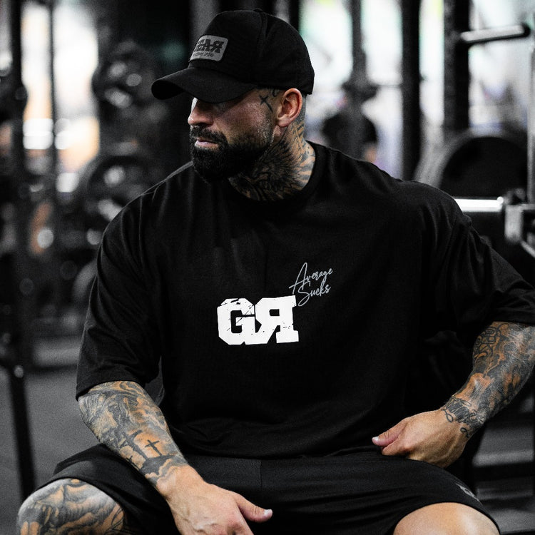 Oversized musta treenipaita salille – GR-logo ja 'Average Sucks' -teksti, miesten urheilupaita rennolla istuvuudella, päällä tatuoidulla miehellä kuntosalilla