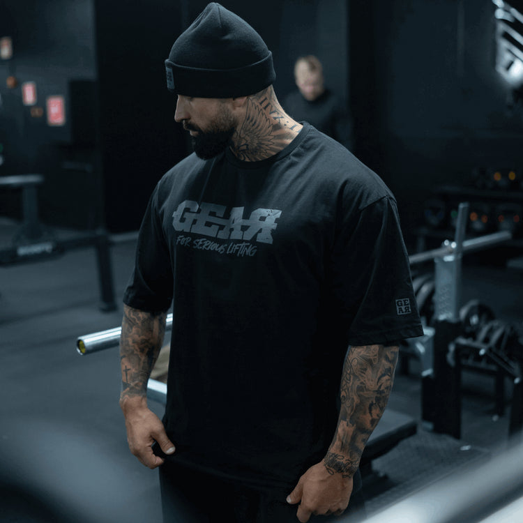 Musta GEAR-treenipaita miehille – 'For Serious Lifting' -slogan rinnassa, tatuoitu kehonrakentaja salilla, rento oversized istuvuus