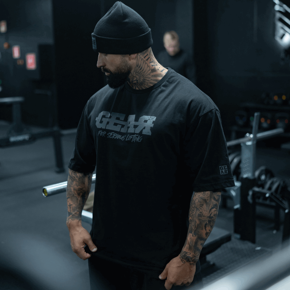 Musta GEAR-treenipaita miehille – 'For Serious Lifting' -slogan rinnassa, tatuoitu kehonrakentaja salilla, rento oversized istuvuus